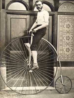 This is an old timey photograph of a man with a mustache riding a penny farthing bicycle.