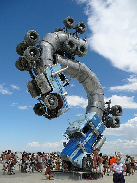 This is a photo of an art installation at Burning Man. It is two truck cabs, one in the air, connected by a large curved steel form.