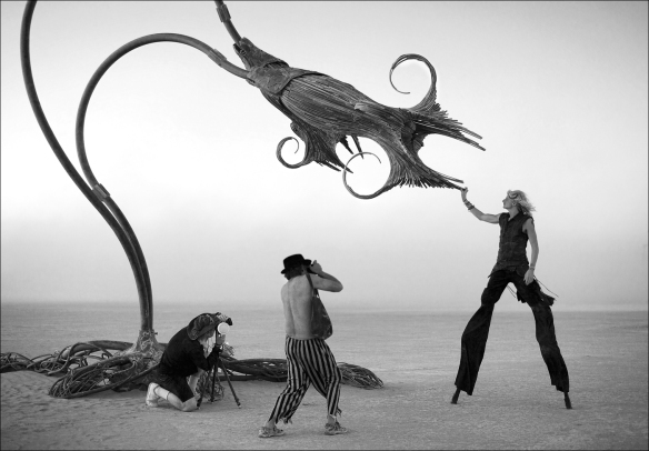 This is an image of a flower-like sculpture art installation at Burning man. There are two men photographing a woman on stilts who is reaching up to touch the sculpture.