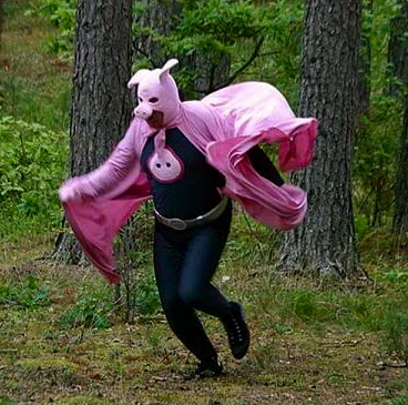 This is an image of a man in a spandex bodysuit, wearing a pig mask attached to a pink cape and running through the forest